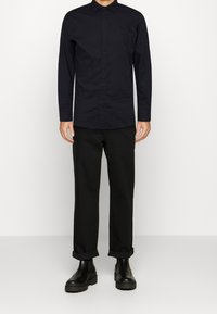 Černá košile s dlouhým rukávem na knoflíky, vyrobená z bavlny, v kombinaci s černými ohrnutými kalhotami a masivními černými kotníkovými botami.