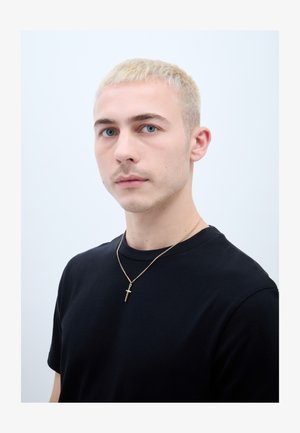 Junger Mann mit kurzen blonden Haaren, blauen Augen, trägt ein schwarzes Hemd und eine goldene Kreuzkette, blickt nach vorn mit neutralem Gesichtsausdruck.