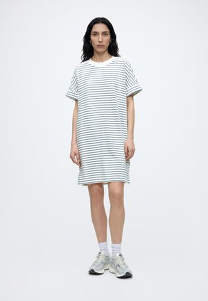 Kobieta z długimi ciemnymi włosami, ubrana w białą sukienkę z cienkimi poziomymi granatowymi paskami, białe skarpetki i białe trampki, stoi na jednolitym tle.