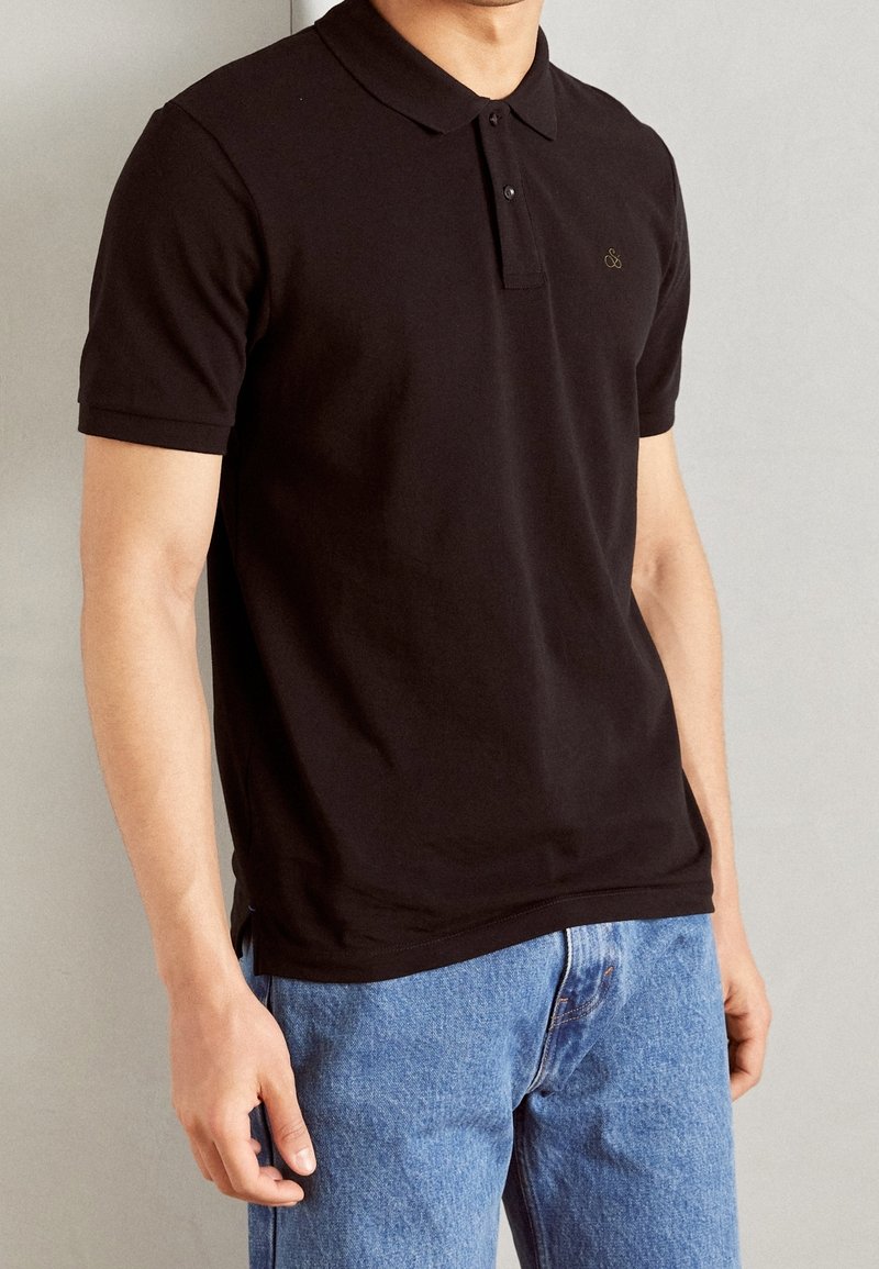 Polo en coton noir avec col et fermeture à trois boutons, associé à un jean bleu clair. Doté d'un petit logo sur la poitrine.