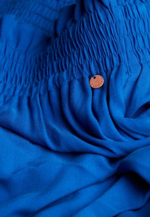 Blauer Stoff mit geraffter Textur und glatter Oberfläche, akzentuiert durch ein kleines, rundes Etikett mit eingeprägtem Markennamen.