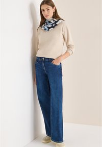 Béžový pletený svetr, tmavě modré džíny s širokými nohavicemi a předními kapsami, a světle zbarvené tenisky. Šála s modrými abstraktními vzory kolem krku.