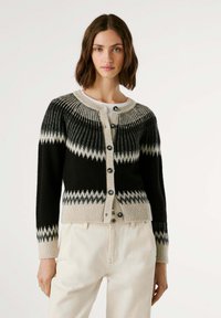 Cardigan negro y crema con patrón de zigzag, escote redondo, textura acanalada y seis botones frontales. Lleva puesto sobre una blusa blanca.