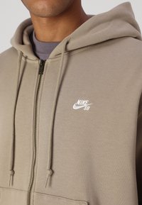 Bežna Nike SB dukserica s patentnim zatvaračem, kapuljačom na vezanje i malim bijelim logom na prsima. Mekani materijal s teksturiranim završetkom.