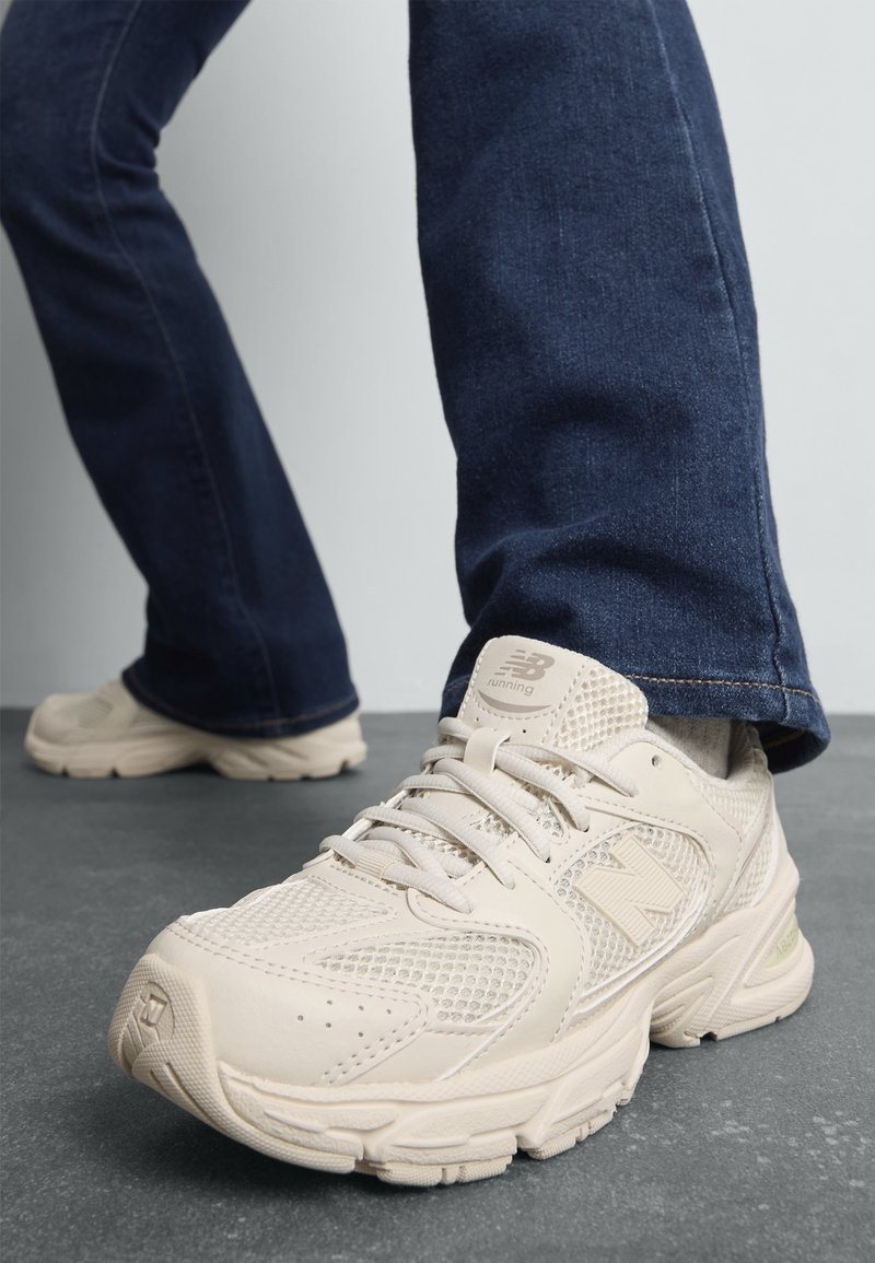 Personne portant un jean bleu et des baskets de course beige New Balance, debout sur un sol gris devant un mur clair.