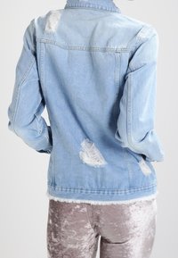 Person i ljusblå, slitna jeansjacka och glansiga, strukturerade, beige sametsbyxor, sedd bakifrån mot en enfärgad bakgrund.