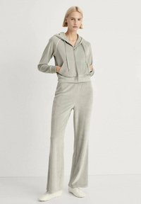 Tuta in velour grigio chiaro, caratterizzato da una felpa con zip e cappuccio con coulisse e tasche frontali, abbinato a pantaloni a zampa d'elefante e sneaker bianche.