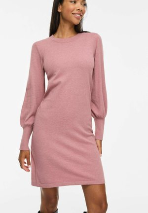 Femme portant une robe en maille rose poudré à manches longues et longueur genou, debout devant un fond blanc uni, souriante et regardant sur le côté.