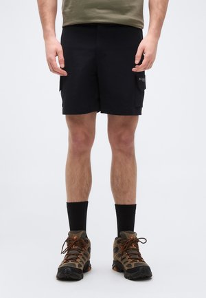 Mand iført sorte cargobukser, sorte crew-sokker, brune og sorte vandrestøvler, stående foran en ensfarvet hvid baggrund.