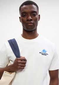 Valge puuvillane T-särk sinise broderitud "Marc O'Polo DENIM" logoga. Mudel hoiab käes beeži kott, olles seljas tumesinine rihm.