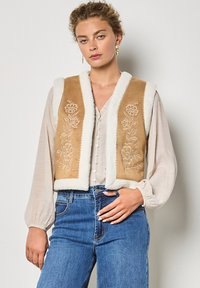 Beige bestickte Weste mit weißem Schaffellbesatz, die mit floralen Mustern versehen ist. Getragen über einer hellen Bluse und kombiniert mit hochtaillierten blauen Jeans.