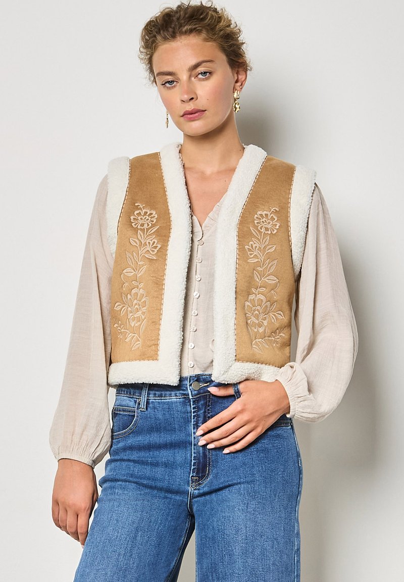 Chaleco bordado en tono canela con borde de piel sintética blanca, que presenta patrones florales. Llevado sobre una blusa clara y combinado con jeans ajustados de cintura alta en azul.