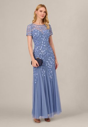 Frau trägt ein bodenlanges lavendelfarbenes Kleid mit Blumenstickerei, hält eine marineblaue Clutch und trägt hängende Ohrringe sowie Sandalen.