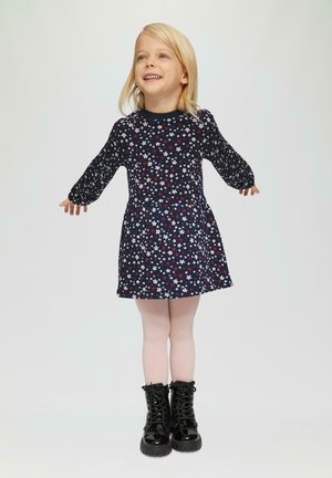 Robe florale marine à manches longues, ornée de fleurs blanches et rouges. Portée avec des collants roses et des bottes noires à lacets.