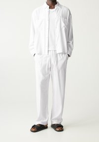 Hvid linnedskjorte med knaplukning, hvid T-shirt under, hvide snøre bukser og sorte slidesandaler med sløjfedetalje.