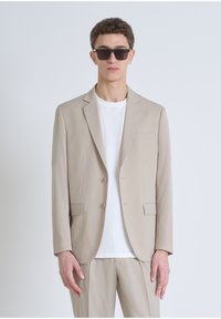 Blazer beige ajusté avec deux boutons à l'avant, revers crantés, poches latérales, porté sur un t-shirt blanc. Les accessoires incluent des lunettes de soleil sombres.