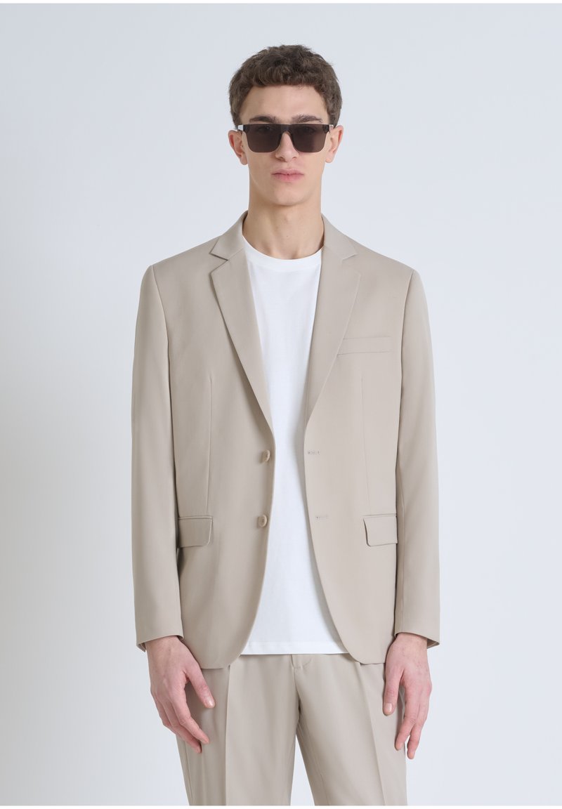 Blazer beige ajusté avec deux boutons à l'avant, revers crantés, poches latérales, porté sur un t-shirt blanc. Les accessoires incluent des lunettes de soleil sombres.