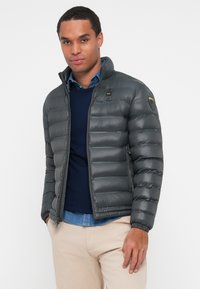 Giacca a vento grigia con finitura lucida, caratterizzata da un colletto alto, chiusura zip, design leggero e polsini elasticizzati, indossata sopra un maglione blu navy.