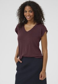 T-shirt bordeaux à col en V avec des manches courtes, texture douce, coupe décontractée, porté avec une jupe bleu foncé, main dans la poche.