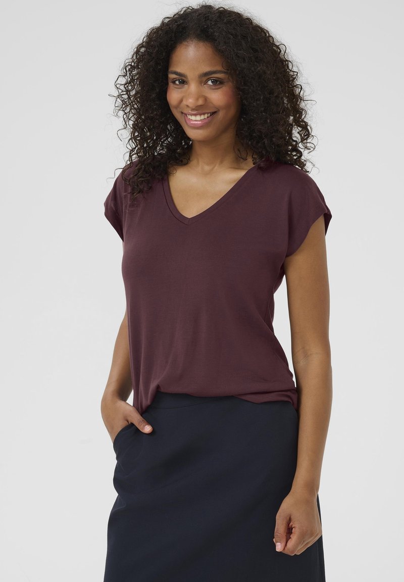 T-shirt bordeaux à col en V avec des manches courtes, texture douce, coupe décontractée, porté avec une jupe bleu foncé, main dans la poche.