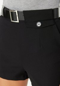 Pantalones cortos negros ajustados con una tela suave, que presentan un cinturón rectangular negro con una hebilla plateada y un cierre de botón en la cintura.