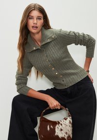 Olivgrüner kurz geschnittener Cardigan mit Rippenstruktur, mit Knopfleiste und weitem Kragen. Kombiniert mit dunklen, weit geschnittenen Hosen und einer braunen, gepunkteten Tasche.