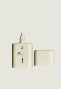 Bouteille en plastique beige de fond de teint naturel transparent avec un couvercle à vis. Texte en bleu : "95 + 100 Fond de Teint Naturel Transparent." Design simple et minimal.