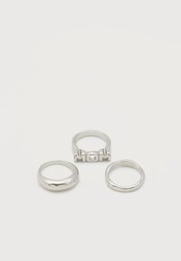 Τρεις ασημένιοι κρίκοι σε λευκή επιφάνεια, ένας απλός βραχίονας, ένας λεπτός βραχίονας, και ένας με ανασηκωμένα γράμματα "MOM" στην κορυφή.