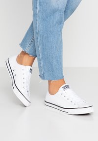 Hvite lerretssneakers har et lavt design, svart gummisåle med hvit kant og seks metalløyer. Brukt sammen med lyseblå denimjeans.