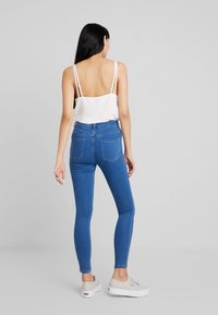 Jeans ajustados azul con cintura alta, con diseño de bolsillo trasero. Combinados con una camiseta sin mangas blanca y zapatillas gris claro.