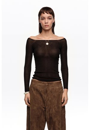 Joven con cabello corto y castaño que lleva una blusa negra transparente sin hombros, pantalones de ante marrones, una pulsera blanca y un collar con colgante en forma de sol.