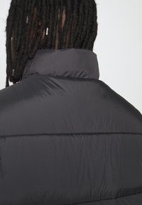 Černá prošívaná bunda s vysokým límcem, s horizontálními švy a hladkou, lesklou texturou. Pohled zezadu zdůrazňuje minimalistický design.