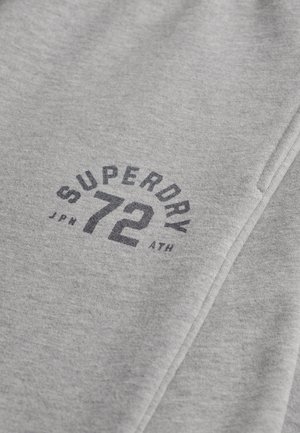 Tissu gris avec "SUPERDRY 72 JPN ATH" imprimé en gris foncé sur la surface, montrant une couture pliée.