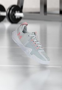 Αθλητικό παπούτσι ανοιχτού γκρι και ροζ που αιωρείται πάνω από το δάπεδο γυμναστηρίου με θολό εξοπλισμό αλτήρων στο φόντο.