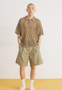 Kortermet brun mønstret polo skjorte med krage, kombinert med lyse olivengrønne plissert shorts; modellen har på seg hvite joggesko.