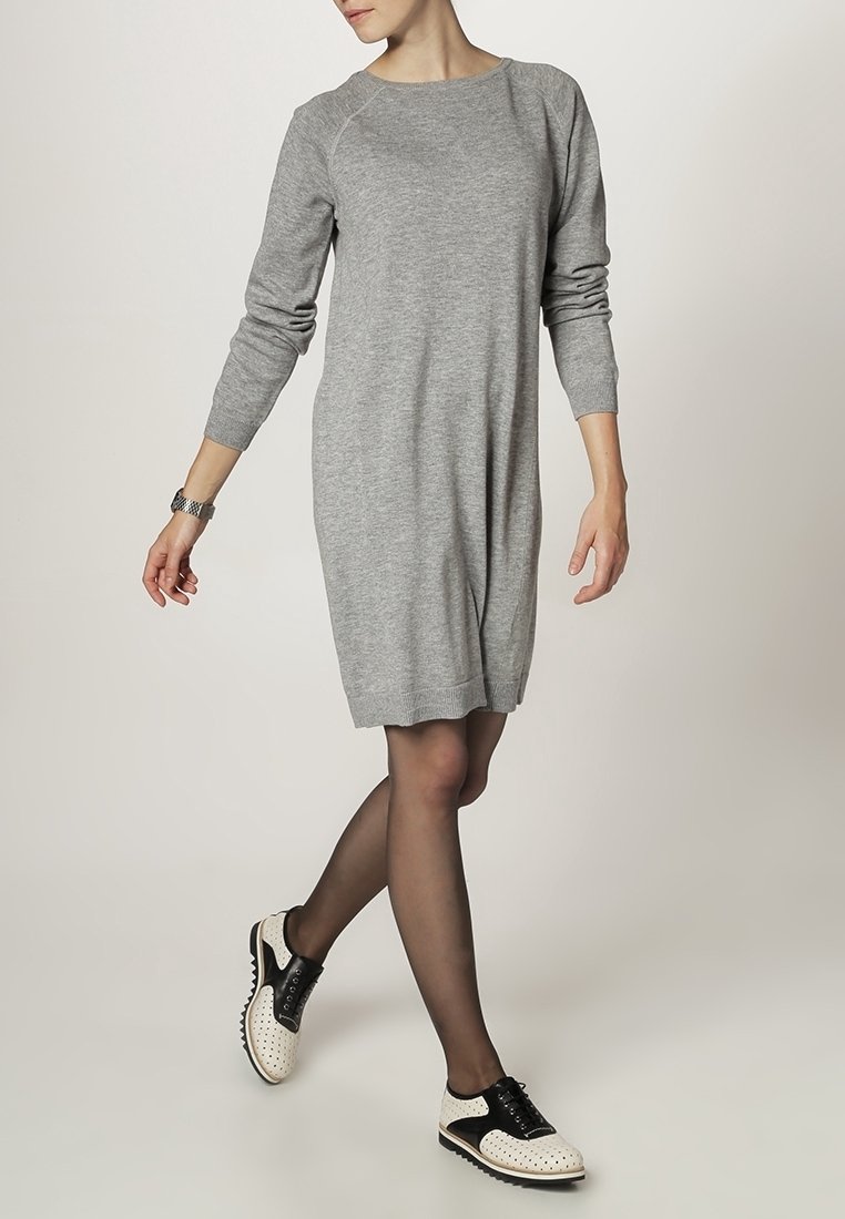 Femme portant une robe en maille grise à manches longues, des collants noirs transparents et des chaussures noires et blanches à lacets, posant avec une jambe croisée vers l'avant.