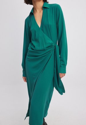 Femme portant une robe portefeuille à manches longues bleu sarcelle avec un détail de nouage sur le côté, un col et un devant drapé sur un fond uni.