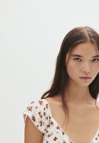 Rochie albă cu mâneci scurte și bufante, având modele florale roșii și verzi. Țesătură cu textură, cu un corp smockat și dantelă la margine.