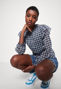 Camicia a quadri con maniche arricciate in blu e bianco, abbinata a pantaloni corti in denim strappato e sneakers blu e bianche.
