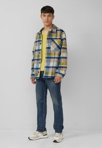 Chemise en flanelle à carreaux bleue, jaune et blanche ; t-shirt jaune à col en V en dessous ; jeans bleus ; baskets gris clair et crème.