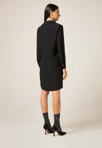 Zwarte jurk met lange mouwen en kraag, met een gladde textuur en knoopmanchetten. Gecombineerd met zwarte sokken met patroon en hoge hakken.