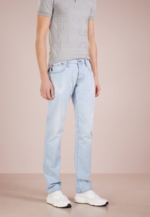 Homme portant un jean bleu clair, un t-shirt gris à manches courtes et des baskets blanches, debout devant un fond neutre.