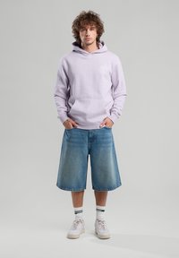 Laventelitakki, jossa on etutasku ja logo, yhdistettyinä polvipitkiin farkkushorteihin. Valkoiset tennarit, joissa on vihreät korostesukat, täydentävät asun.