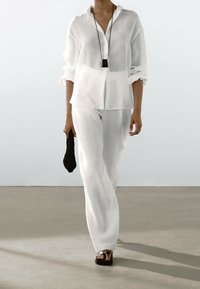 Camicia bianca in lino con maniche arrotolate, abbinata a pantaloni larghi bianchi in lino. Una pochette nera texturizzata e sandali marroni completano l'outfit.