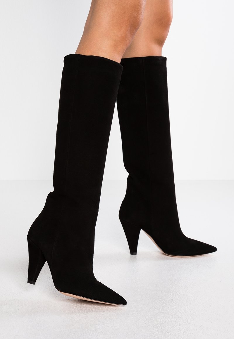 Des jambes portant des bottes à talons hauts en suède noir, à bout pointu et mi-mollet, se tenant sur un sol blanc contre un fond blanc.