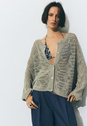 Junge Frau mit kurzen dunklen Haaren, die einen gemusterten Bikini-Top unter einer locker gestrickten beigen Strickjacke und dunklen weiten Hosen trägt, posiert vor einer weißen Wand.