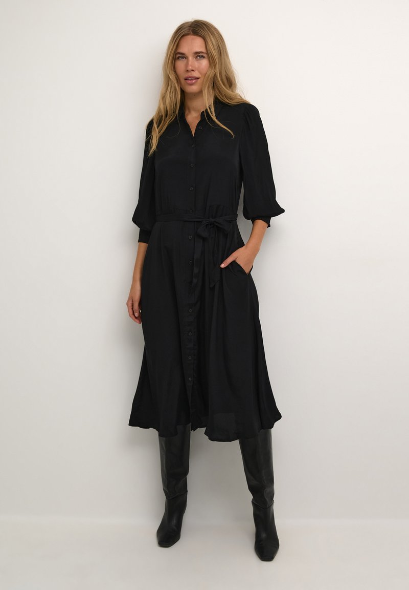 Zwarte midi-jurk met knopenvoorzijde, lange mouwen, een strik taille en zijzakken. Soepele stof met een vloeiende silhouette. Zwarte kniehoge laarzen.