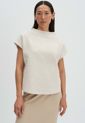 Femme portant un haut texturé écru à manches courtes et une jupe beige, debout devant un fond gris clair uni.