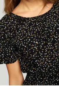 Svart blus med korta ärmar, prydd med ett blommönster av vita och senapsgula blommor på en mörk bakgrund och med smockad midjedetalj.