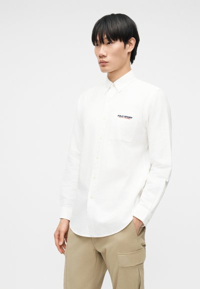 Άνδρας μοντέλο φοράει λευκό πουκάμισο με μακριά μανίκια και κουμπιά, με το λογότυπο "Polo Sport" στην τσέπη του στήθους και μπεζ παντελόνι cargo, κοιτάζοντας προς τα πλάγια.